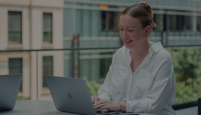 Kvinna i vit skjorta sitter framför en laptop. Kvinnan ler och tittar ner på skärmen.